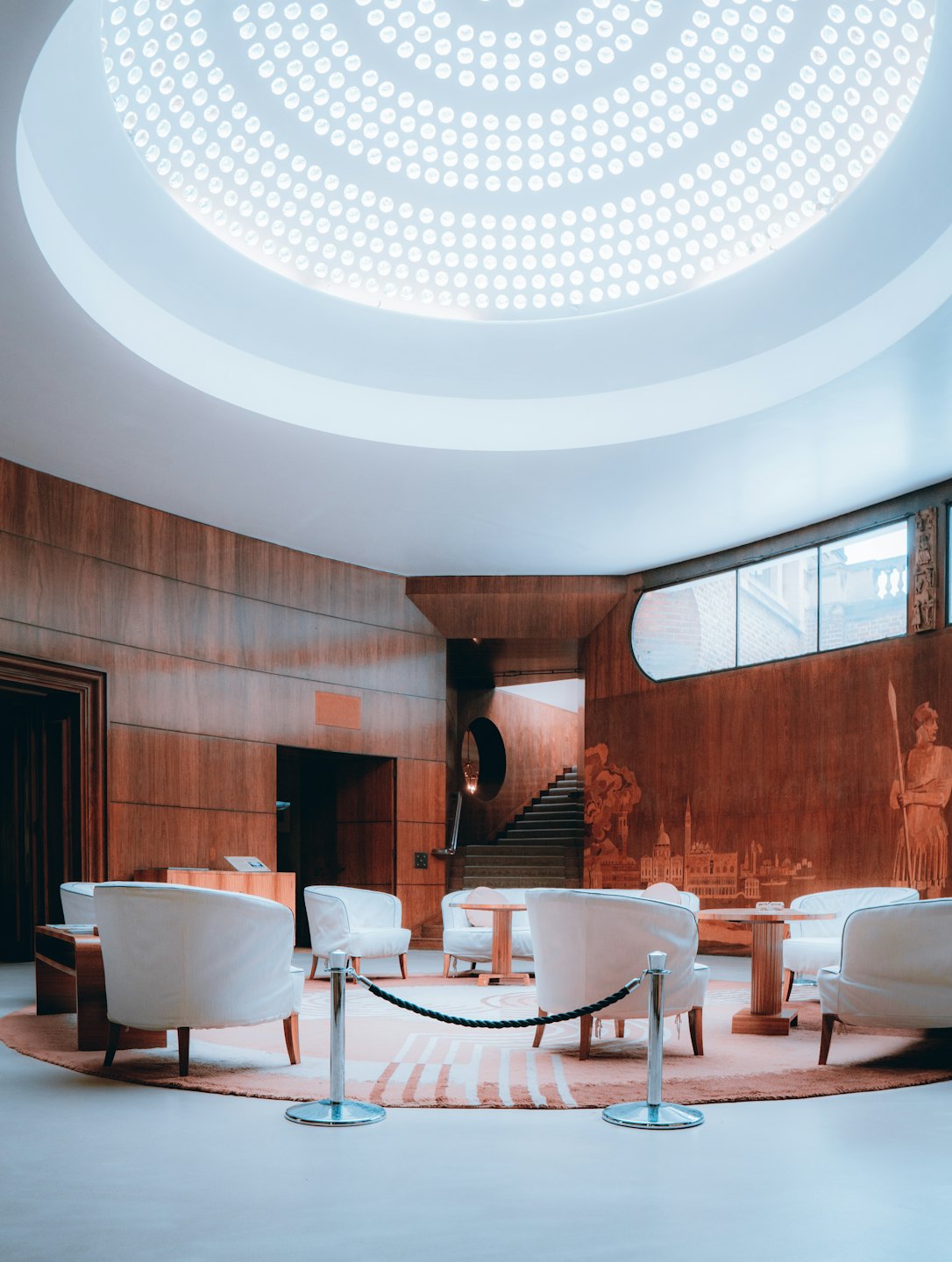 Lobby moderno con tragaluz circular, sillas blancas y mesas de madera.